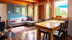 Una habitación acogedora con techo de madera, un sofá gris, una mesa de comedor con sillas y un ventanal con un paraguas azul.