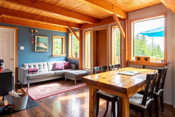 A cozy room with a wooden ceiling, a gray sofa, a dining table with chairs, and a large window with a blue umbrella.