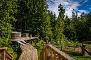 A wooden deck with a hot tub, surrounded by dense forest and a clear sky.