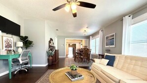 Un salon avec un ventilateur de plafond, une table basse ornée d’une plante, un canapé, un bureau avec une chaise, et un coin repas à l’arrière-plan.
