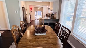Une salle à manger avec une table et des chaises en bois, un lustre, et une vue sur la cuisine et le salon.