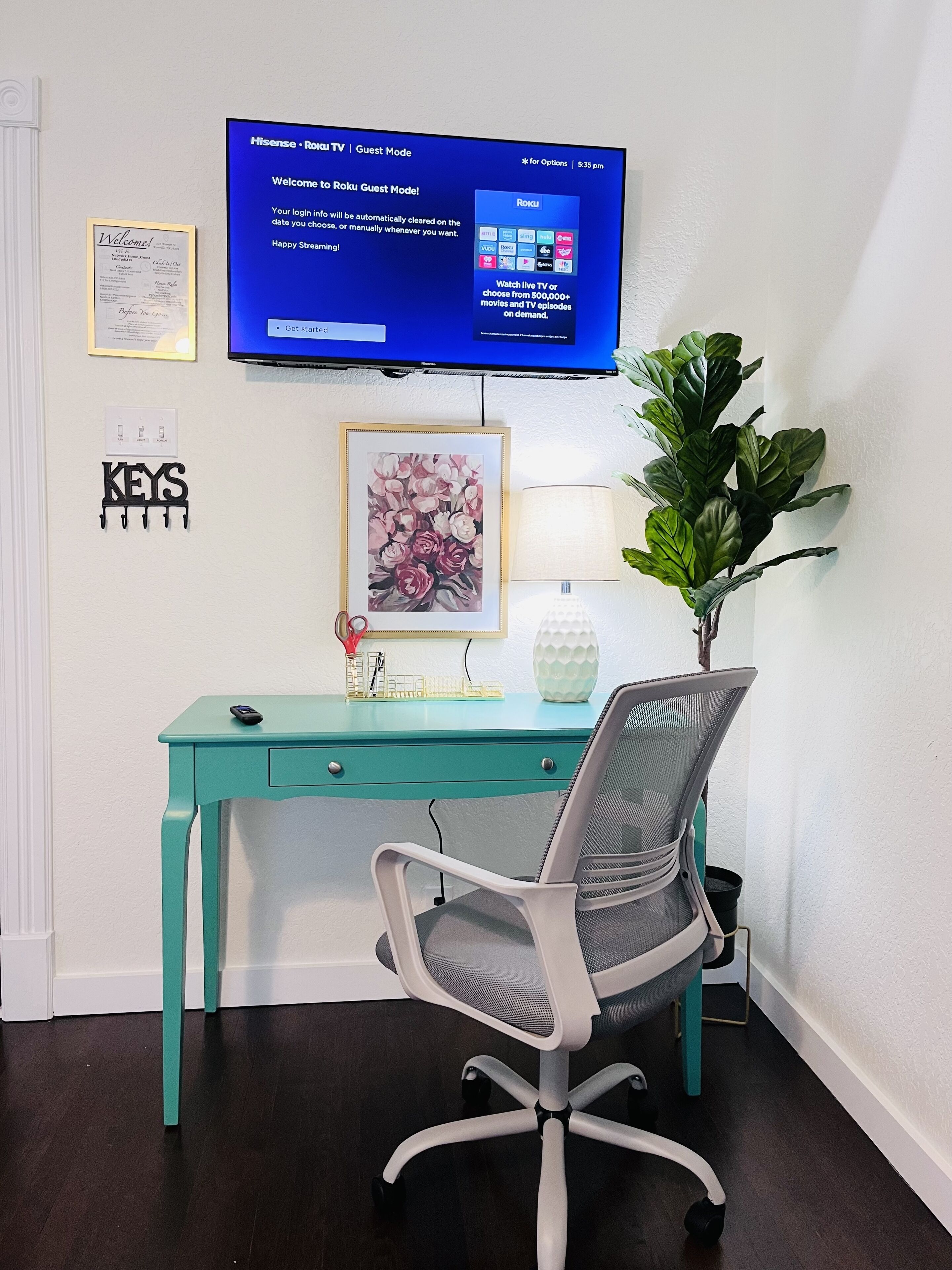 Une pièce avec un bureau de couleur turquoise, une chaise de bureau blanche, une œuvre d’art florale encadrée, une plante en pot, un téléviseur fixé au mur affichant le mode invité Roku et un interrupteur fixé au mur.