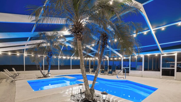 A swimming pool with lounge chairs and palm trees, surrounded by a modern building with large windows and string lights.