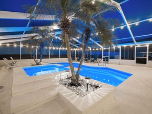 Piscina con sillas de descanso y palmeras, rodeada por un edificio moderno con ventanas amplias y luces colgantes.