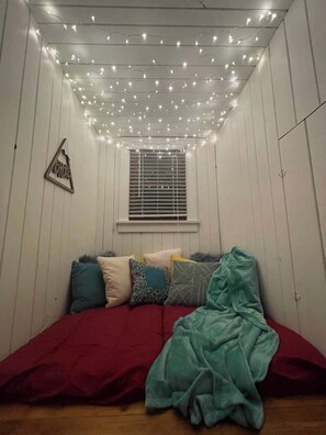 A small, cozy room with a bed, a window with blinds, and string lights on the ceiling.