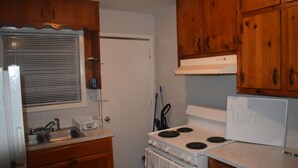 Une petite cuisine avec des meubles en bois, un fourneau blanc, un évier et une fenêtre avec des stores.