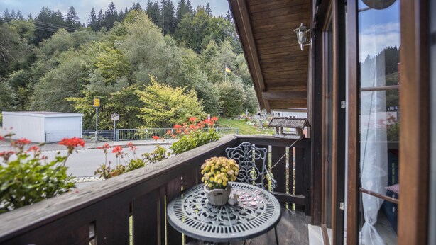 Un balcon en bois avec une table et des chaises, un pot de fleurs et une vue sur une zone boisée.