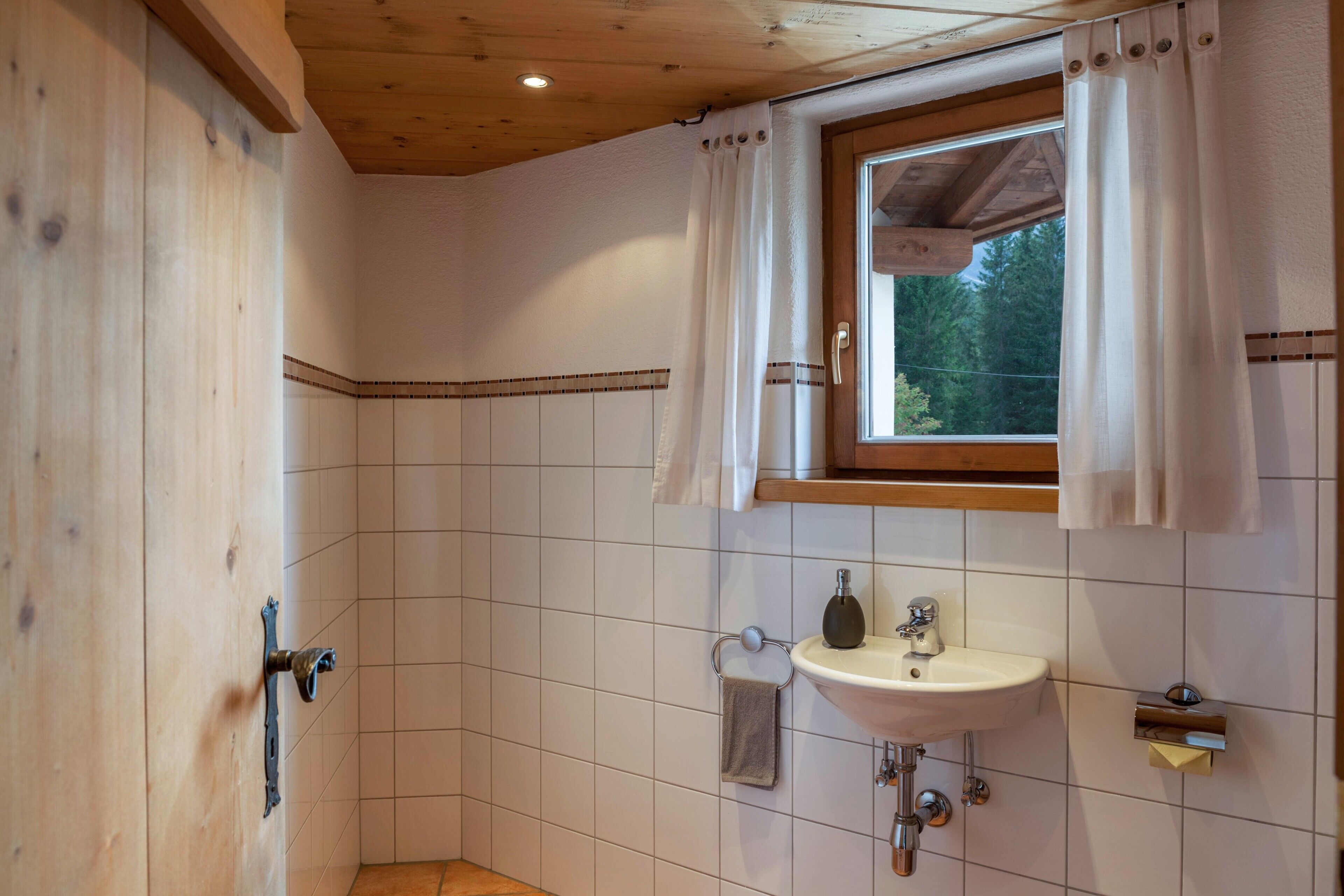Ein Badezimmer mit weiß gefliesten Wänden, einer Holzboden und einem Fenster mit Vorhängen, einem Waschbecken und einem Handtuchhalter.