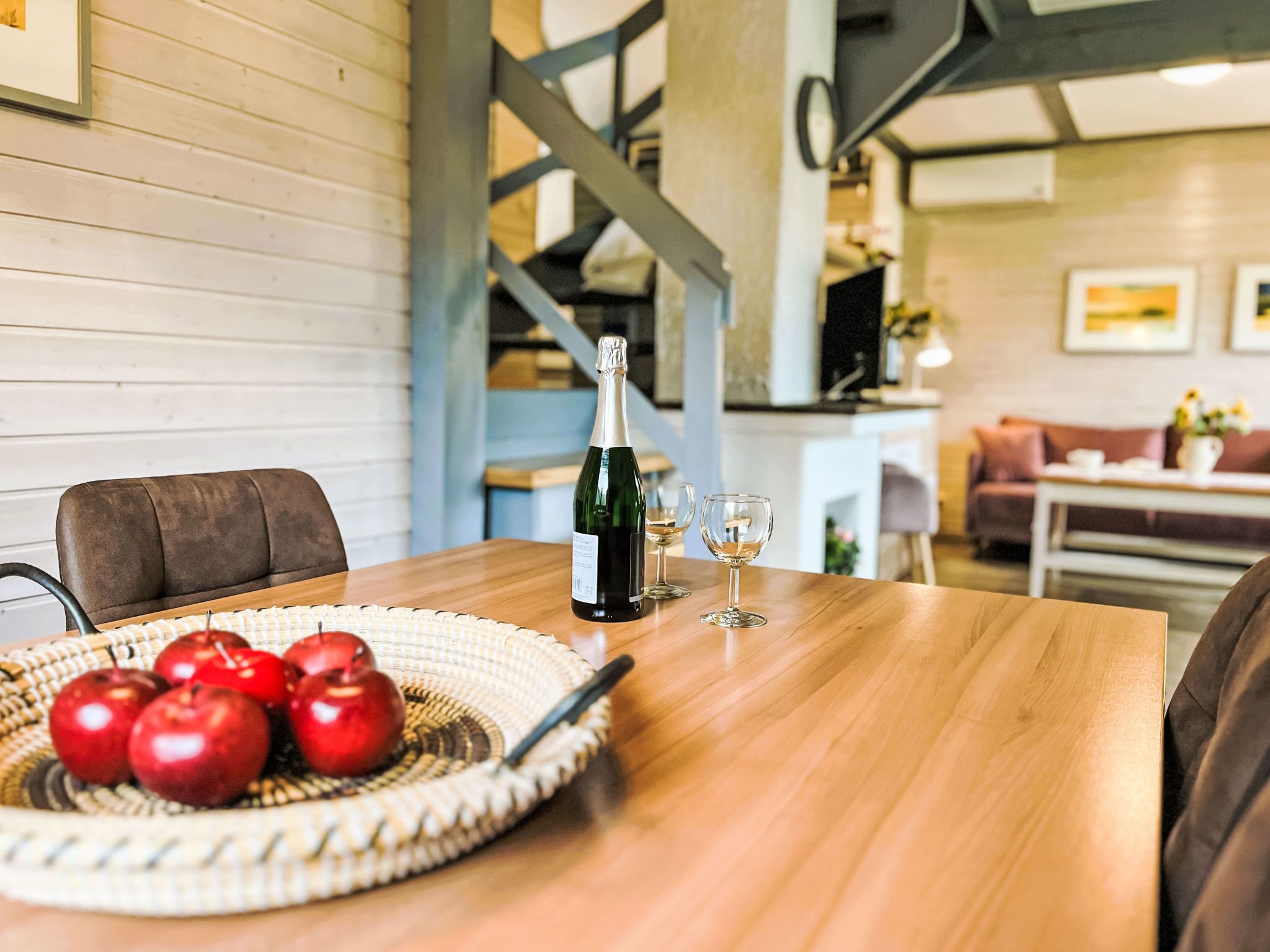 A wooden table with a bottle of champagne, two glasses, and a woven basket of red apples.