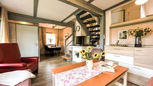 A cozy living room with a wooden table, a red armchair, a staircase, and a kitchen area with a vase of sunflowers.