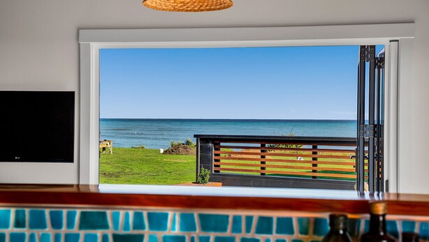 A view from a room with a large window overlooking a beach, a wooden deck, and a tiled backsplash. - Coastal Daze - New Plymouth Holiday Home (New Plymouth)