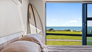 A bedroom with a bed, a surfboard on the wall, and a view of the ocean through an open door. - Coastal Daze - New Plymouth Holiday Home (New Plymouth)
