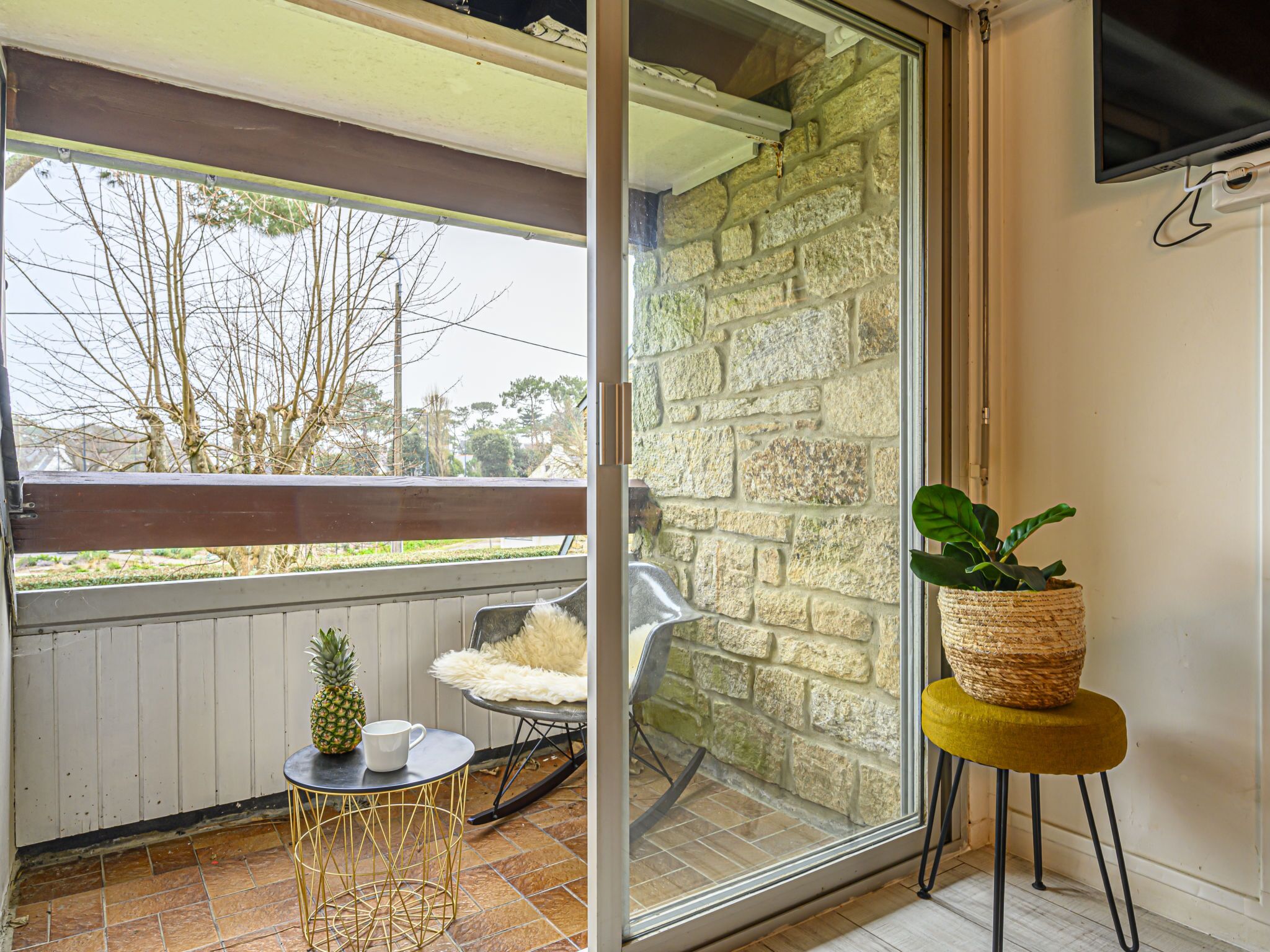 A balcony with a stone wall, a small table with a pineapple and a mug, a rocking chair, and a potted plant on a yellow stool.