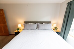 Un lit bien fait, avec du linge de lit blanc, deux tables de chevet avec des lampes et de petites plantes en pot, une porte en bois et une fenêtre avec des rideaux.