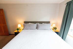 A neatly made bed with white linens, two bedside tables with lamps and small potted plants, a wooden door, and a window with curtains.