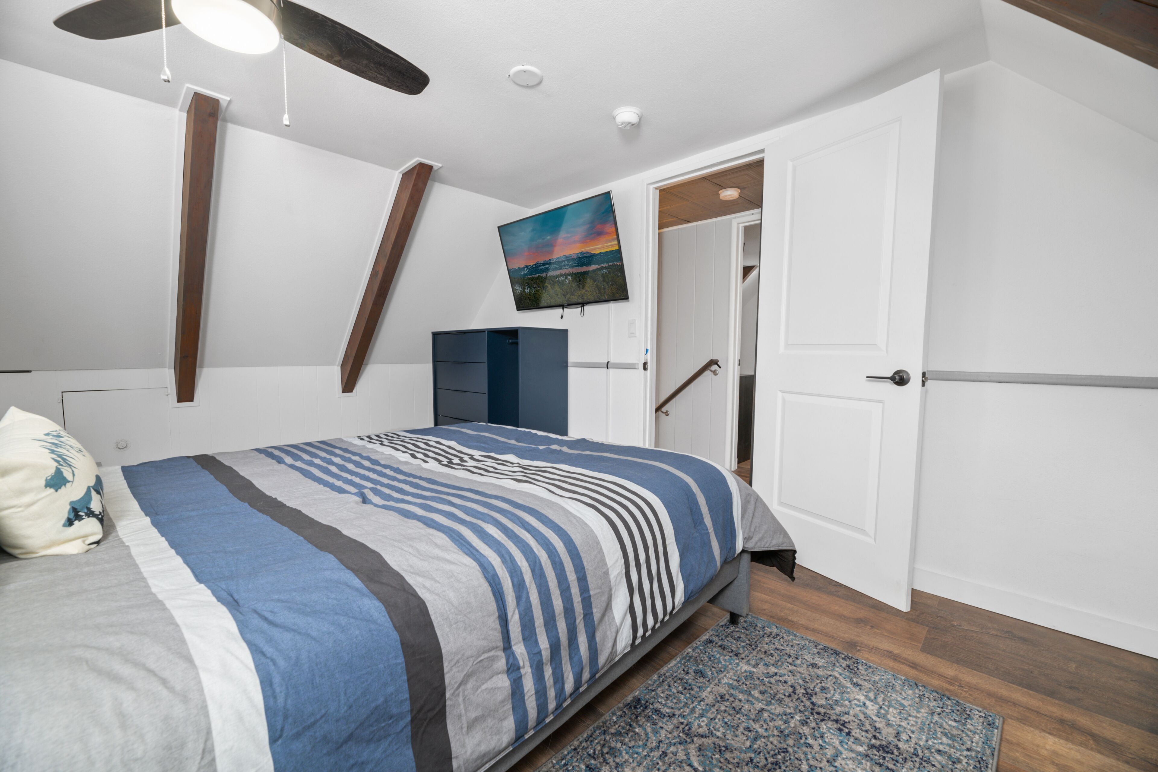A bedroom with a bed, a TV mounted on the wall, a ceiling fan, and a door leading to another room.