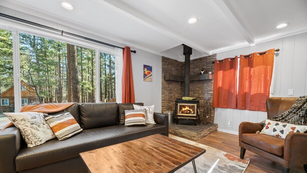 A modern living room with a fireplace, leather sofa, and a large window overlooking a forest. - Vanderhut Chalet - Smart TV, BBQ, Fenced Yard (Sugarloaf)