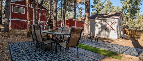 En overdækket terrasse med et mønstret tæppe, et spisebord med stole, og en rød bygning i baggrunden.