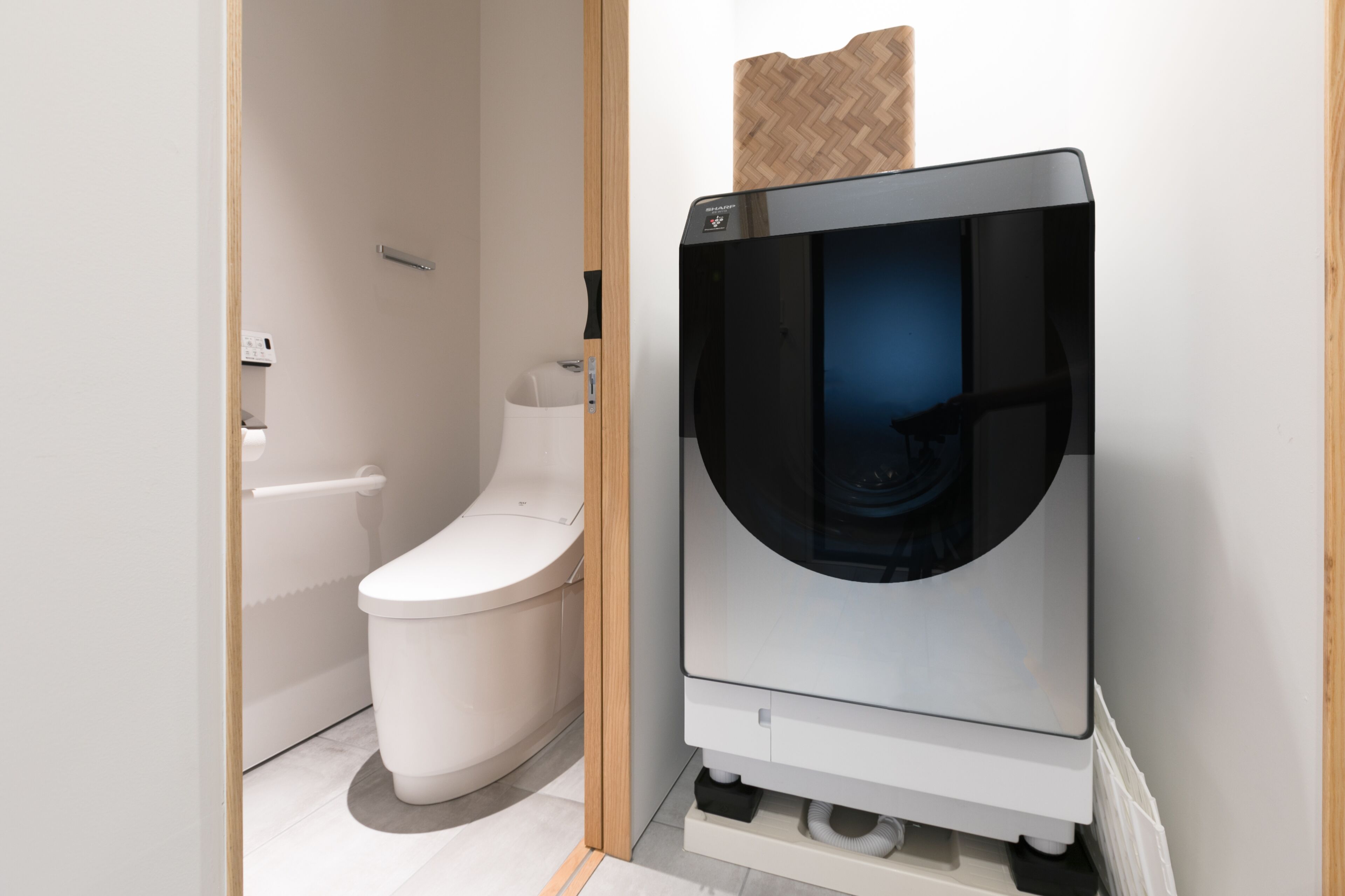 A modern bathroom with a toilet, a wall-mounted washing machine, and a wooden panel above the machine.