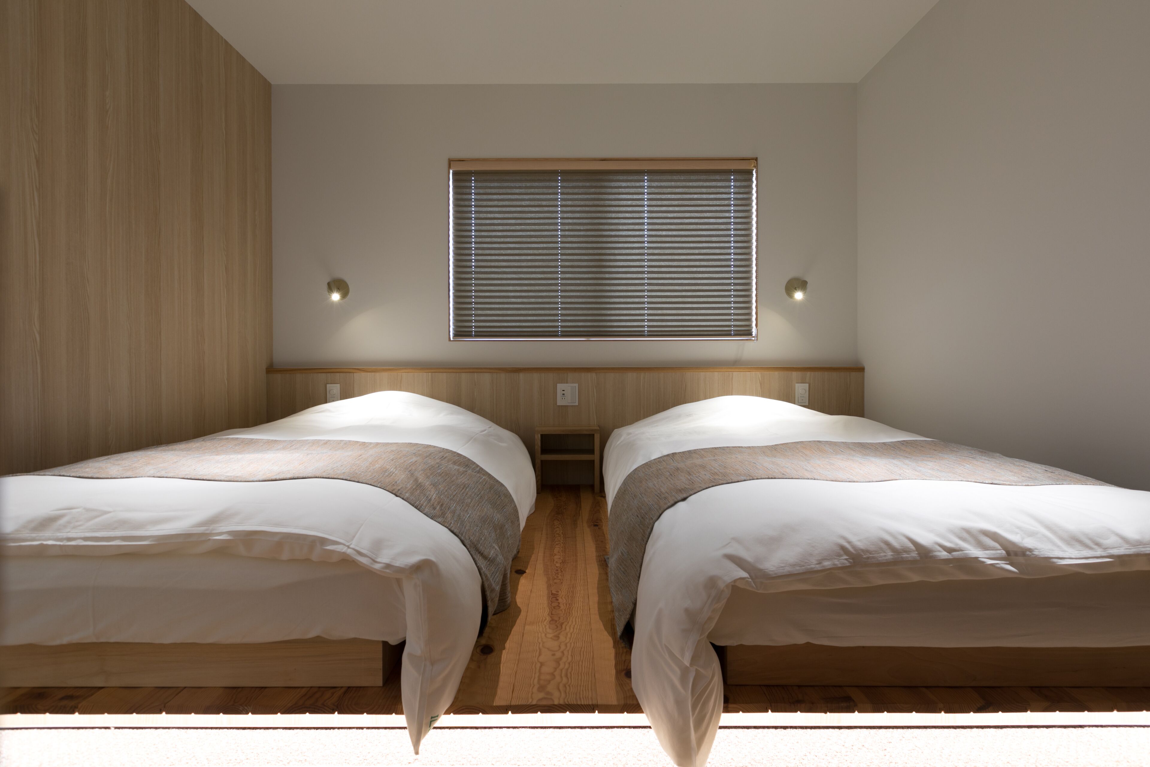 A room with two beds, wooden flooring, a window with blinds, and wall-mounted lights.