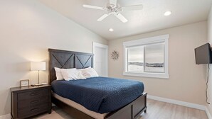 Un dormitorio con cama, mesita de noche, ventilador de techo, ventana con vistas y televisor montado en la pared.