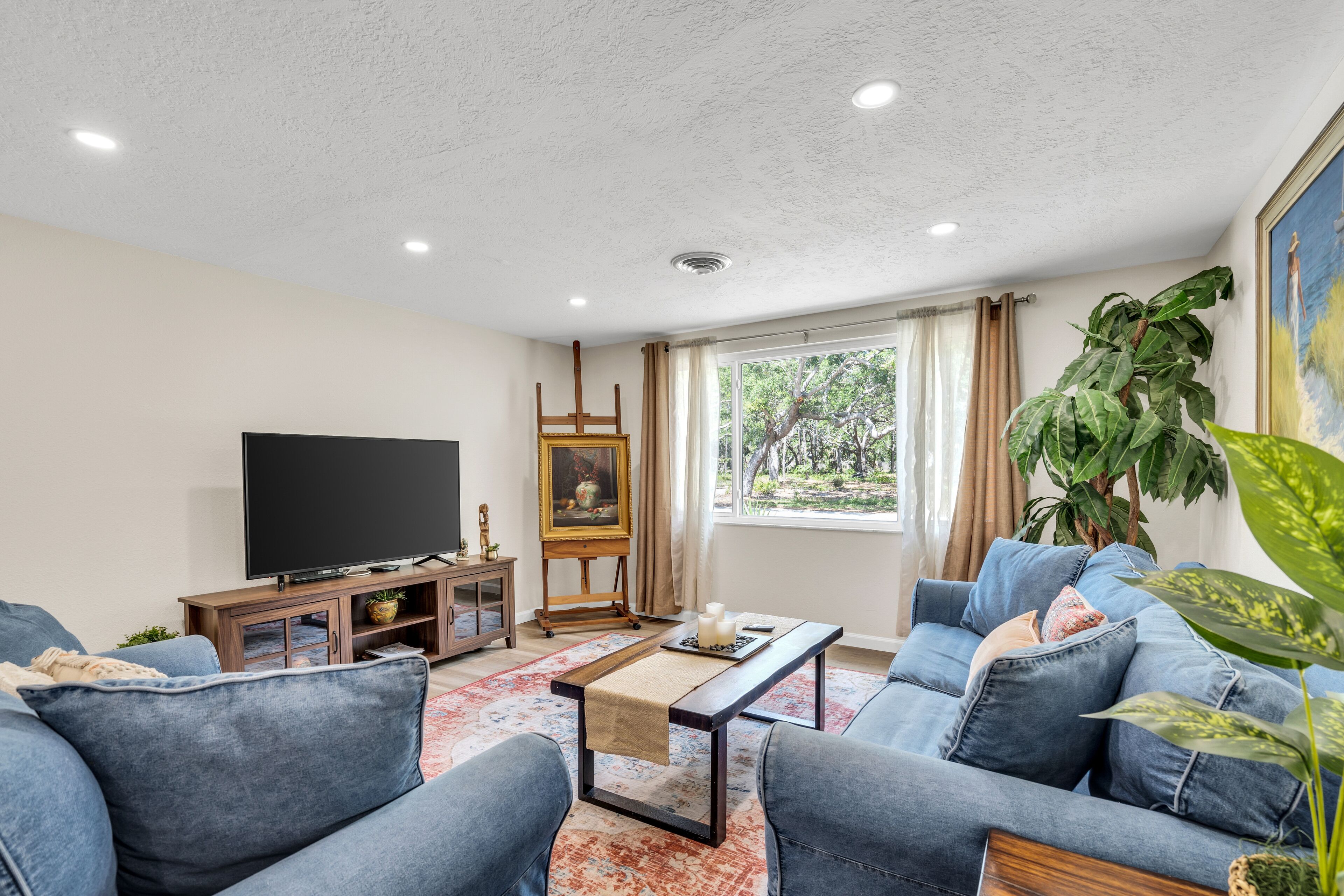 A living room with a flat-screen TV, a wooden cabinet, a painting on easels, a sofa, a coffee table, and a large plant.