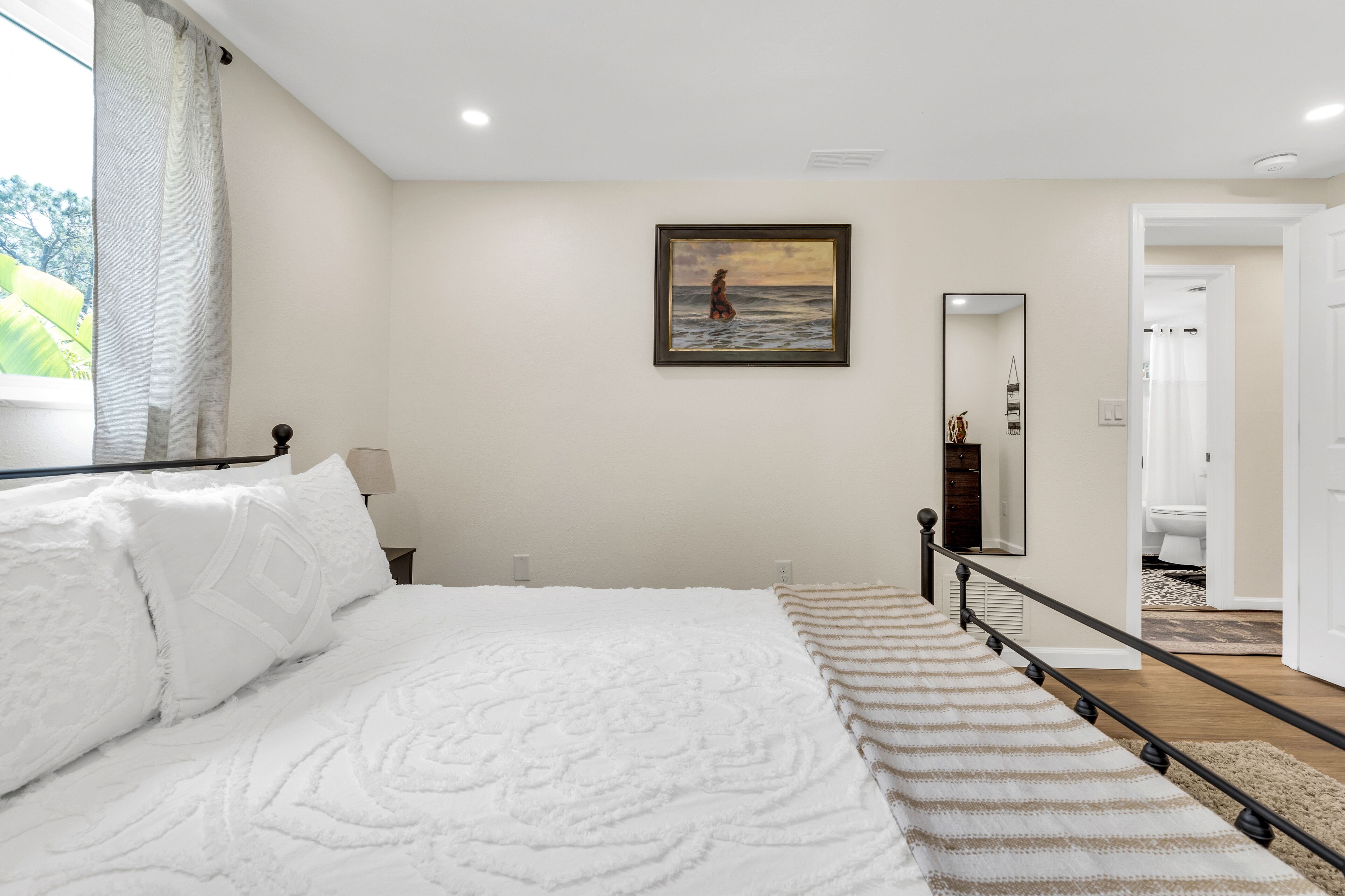 A bedroom with a bed, a painting, a mirror, and a view of the bathroom.