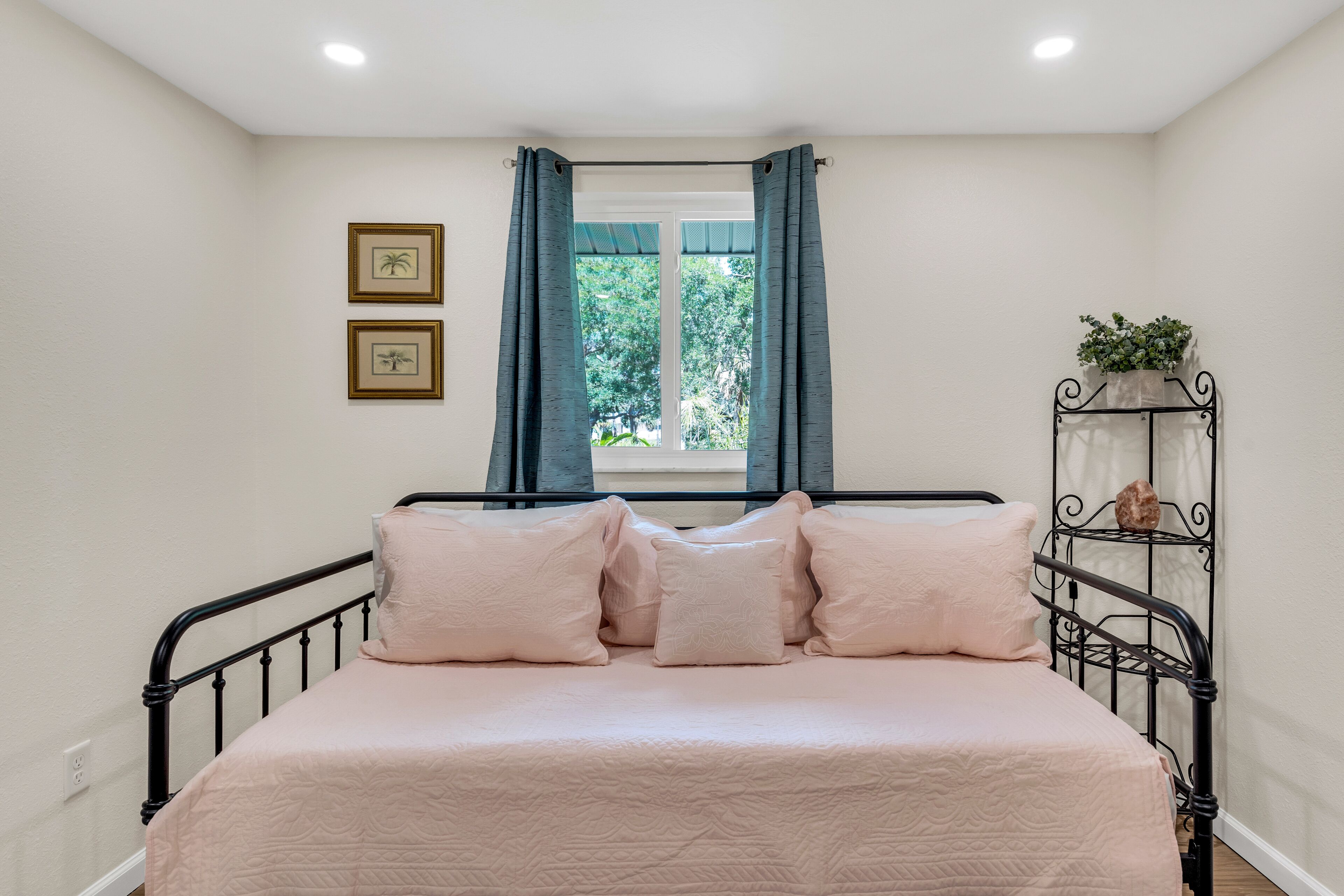 Un lit bien fait, avec une couvre-lit rose et des oreillers, une fenêtre avec des rideaux et une étagère fixée au mur, sur laquelle est posée une plante.