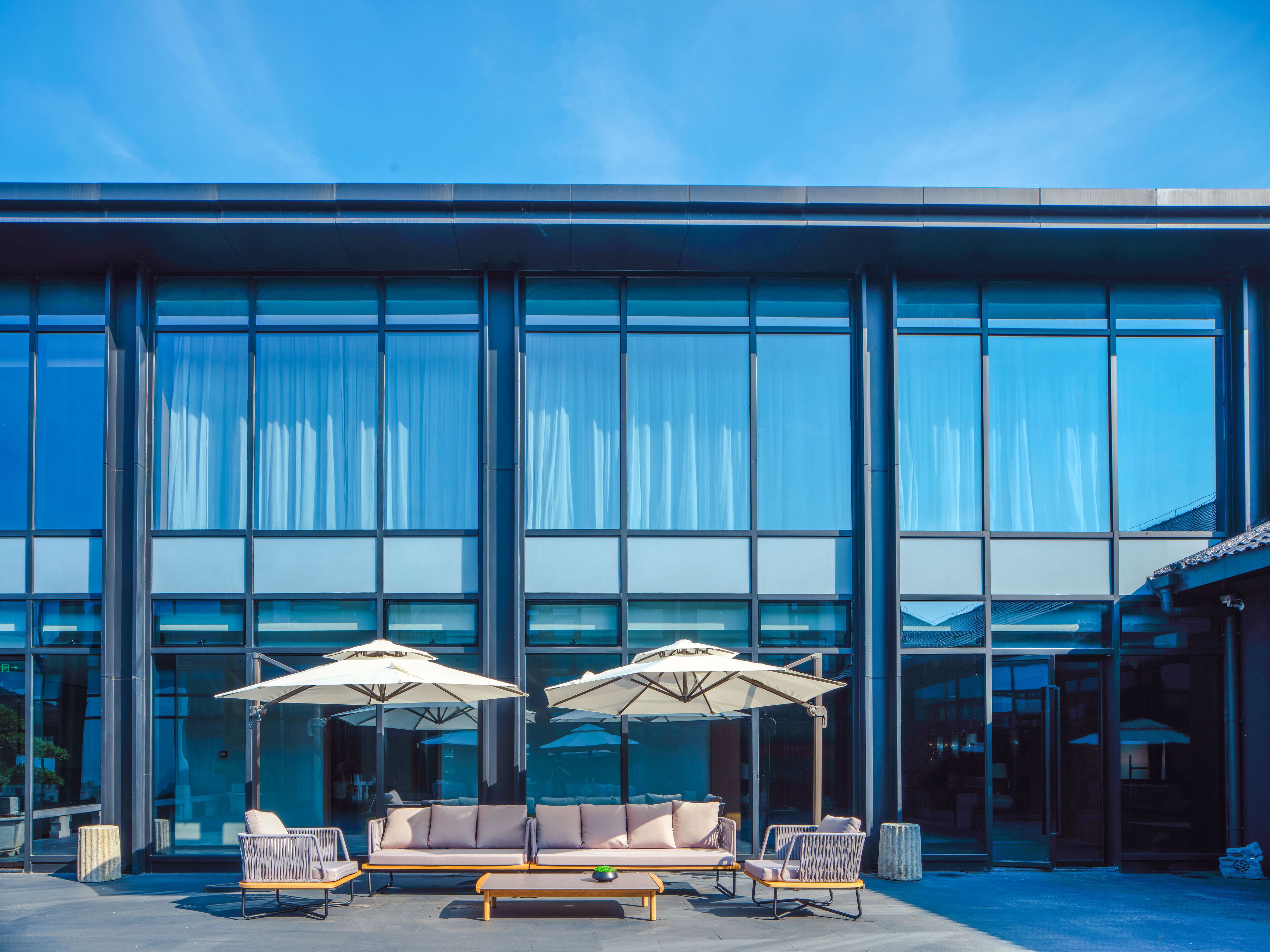 Un bâtiment moderne doté de grandes fenêtres en verre et d’un espace détente avec deux parasols blancs et des meubles en osier.