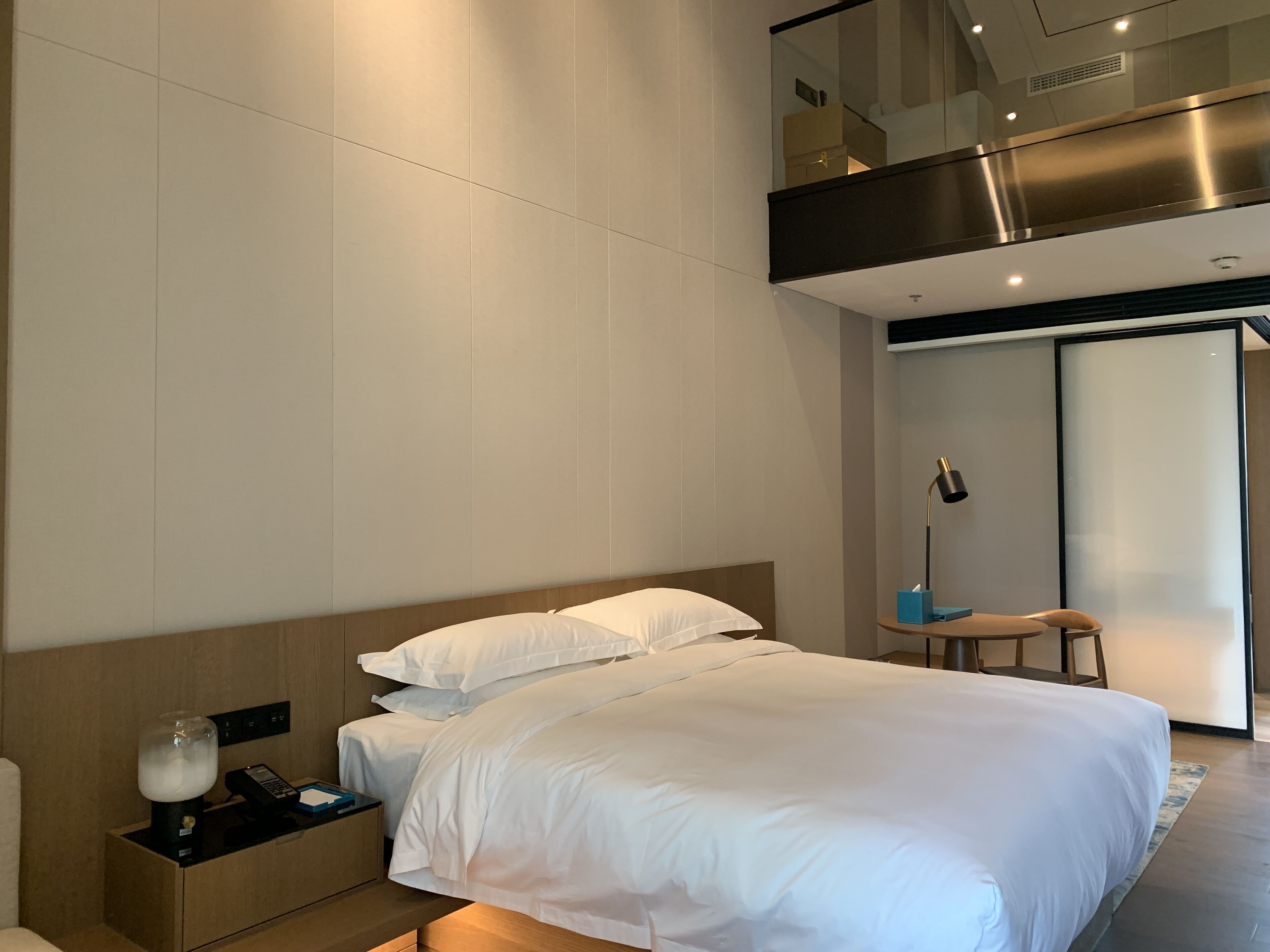 A modern hotel room with a large bed, a bedside table with a lamp, and a small table with a phone and a tissue box.