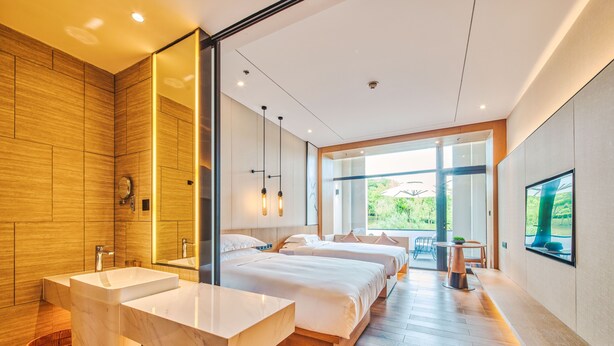 Una habitación de hotel moderna con una cama grande, dos mesitas de noche, un espejo amplio y un baño con bañera, además de vistas al exterior.