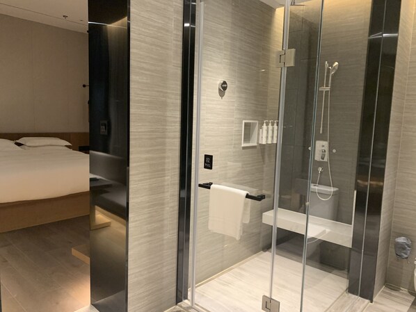A modern hotel room with a glass-enclosed shower, a neatly made bed, and a small bedside table with toiletries.