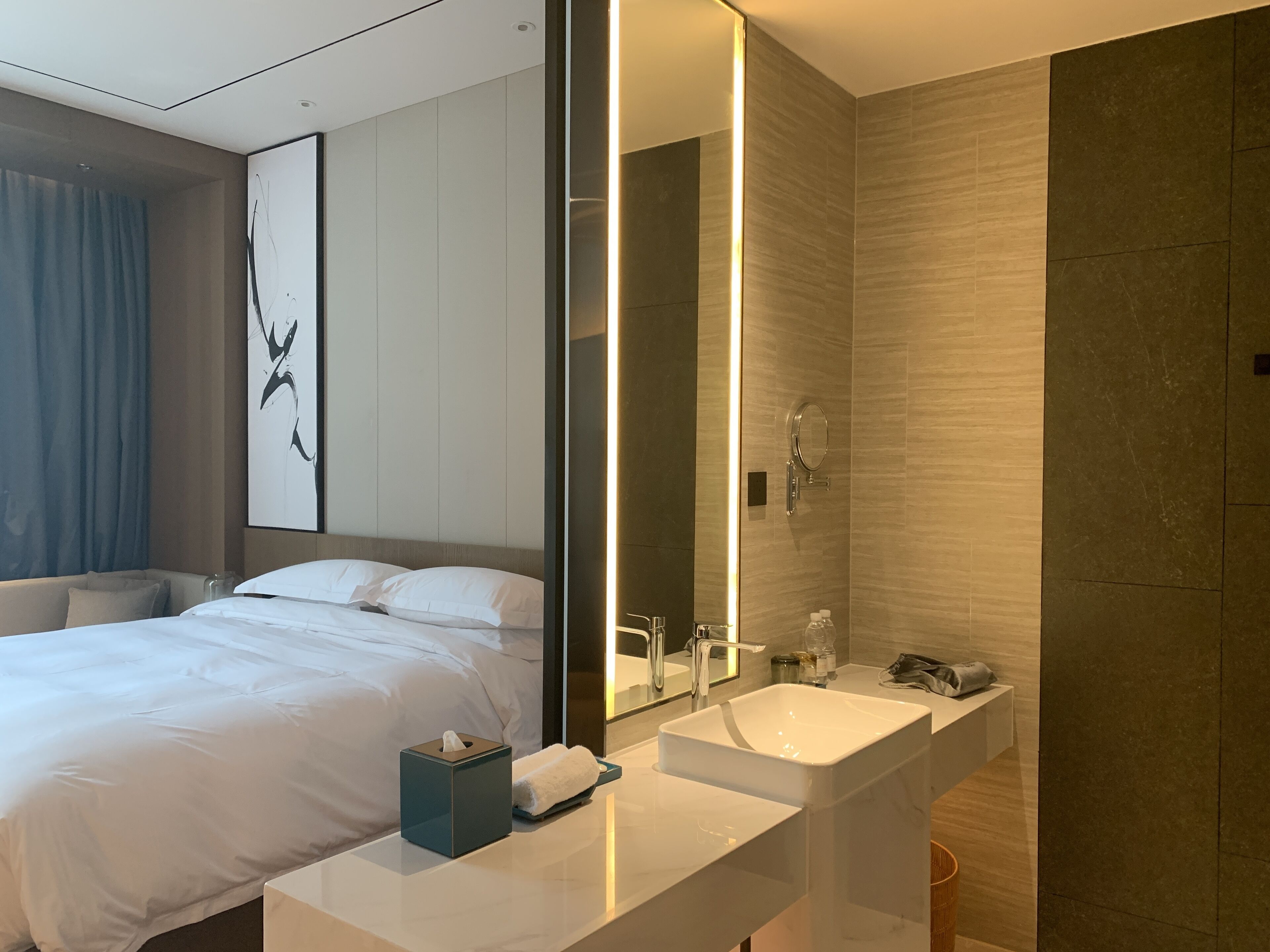 A modern hotel room with a large bed, a bedside table with a tissue box and a towel, a large mirror, and a bathroom with a sink and shower.
