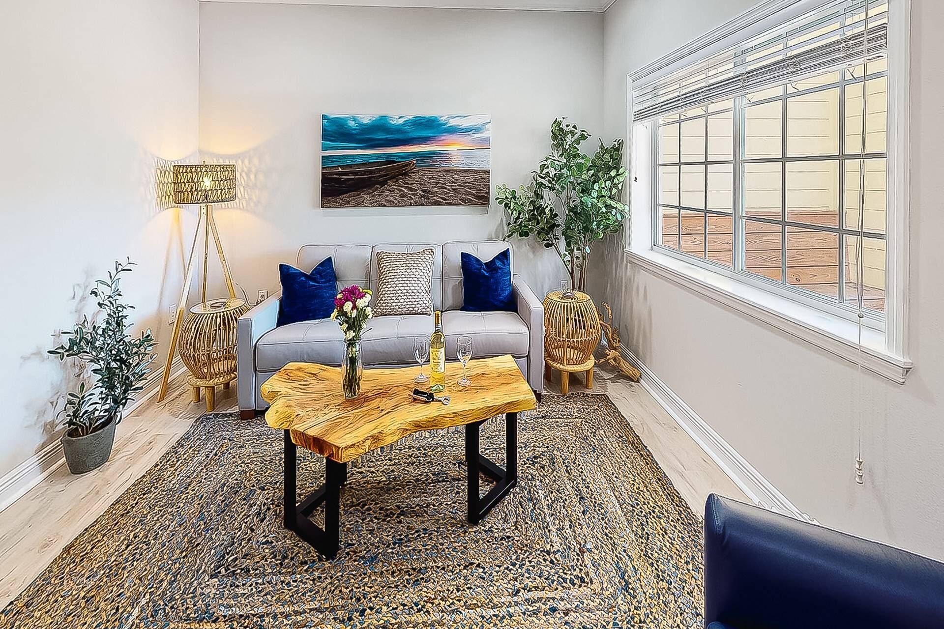 Una sala de estar con sofá, mesa de centro y almohadones decorativos. Hay un cuadro en la pared y una ventana con persianas.