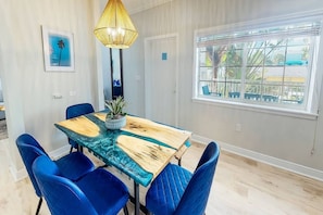 A dining area with a wooden table and blue chairs, a hanging light fixture, and a window with a view of palm trees. - Beach Club | Crescent Beach | Pool & Hot Tub (Sarasota)