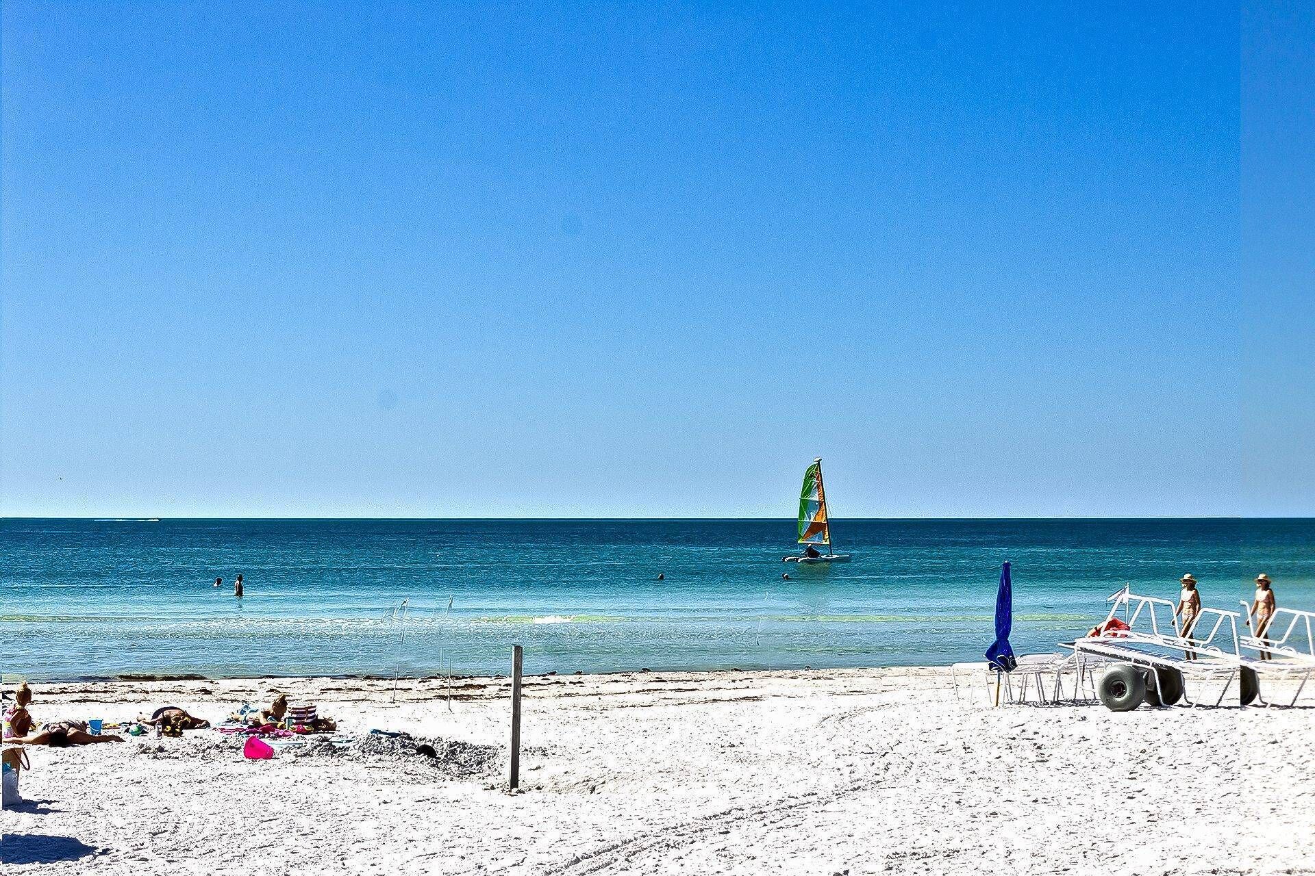 Een strand met een heldere lucht, een zeilboot op het water, en strandstoelen en parasols in het zand.
