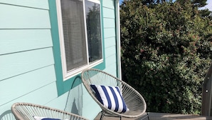 Two wicker chairs with striped cushions on a wooden deck, a teal house with a window, and a bush in the background. - CLEAN Beach Cottage + Beach Path! (Grayland)