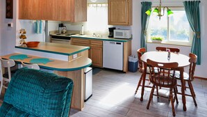 A compact kitchen with wooden cabinets, a white dishwasher, and a dining area with a round table and chairs. - CLEAN Beach Cottage + Beach Path! (Grayland)