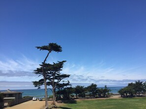 Uitzicht op de kust, met een heldere lucht, een zandstrand met enkele bomen en een gebouw met balkon dat uitkijkt over de oceaan.
