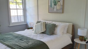 Ein Schlafzimmer mit einem großen Bett, einem Fenster, einem Nachttisch mit Lampe und einem Bild an der Wand.