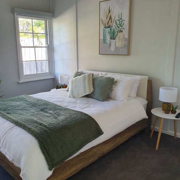 A bedroom with a large bed, a window, a bedside table with a lamp, and a painting on the wall.