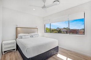 A modern bedroom with a large bed, a ceiling fan, a window with a view of buildings and greenery, and a white bedside table. - TARA5- UNWIND IN LUXURY (Mooloolaba)