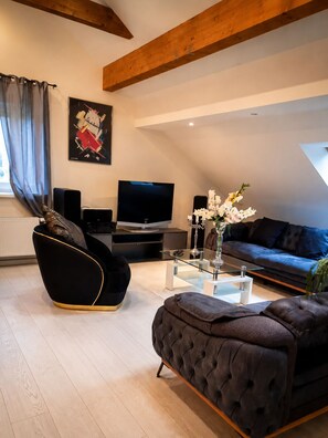 A modern living room with a flat-screen TV, black and gold furniture, a glass coffee table, and a blue sofa. - A peaceful natural oasis with woods and a stream—near Basel, Colmar, and Europa-Park (Basel)