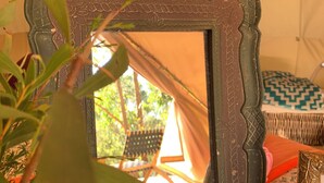 Un miroir rustique doté d’un cadre texturé, une petite plante au premier plan et, à l’arrière-plan, on aperçoit une tente et un coin salon extérieur.