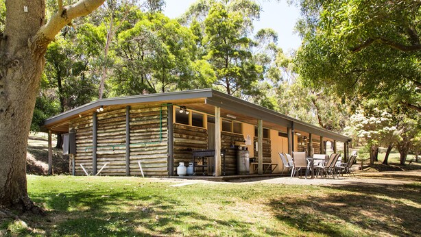 Une cabane rustique en rondins de bois, dotée d’un patio couvert, de coins salon extérieurs et d’un grand arbre au premier plan.