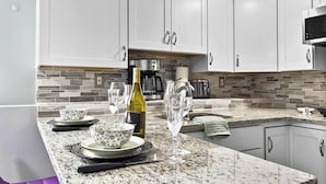 A kitchen with white cabinets, a granite countertop, a coffee maker, and a wine bottle. - Suite at House of L (Chelsea)