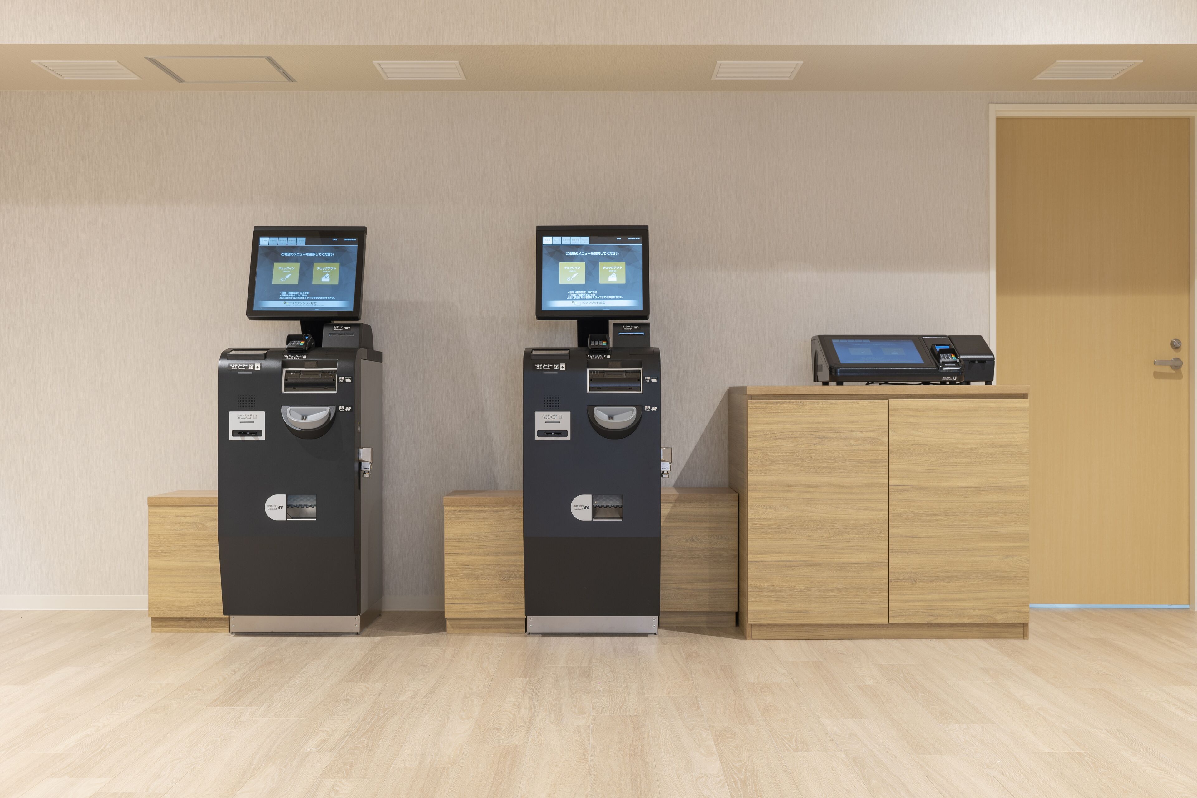 Two self-service kiosks with digital displays, a wooden cabinet, and a door in a room with light-colored flooring.