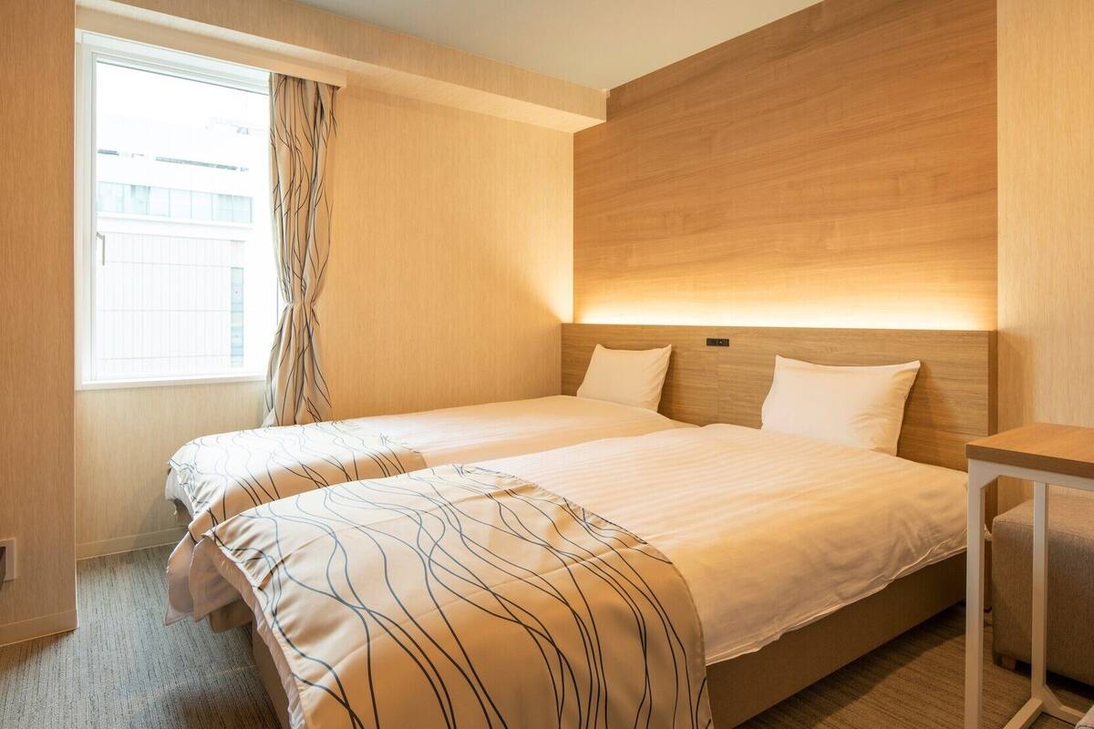 Une chambre d’hôtel avec deux lits, une fenêtre avec des rideaux, une table de chevet et une tête de lit en bois.