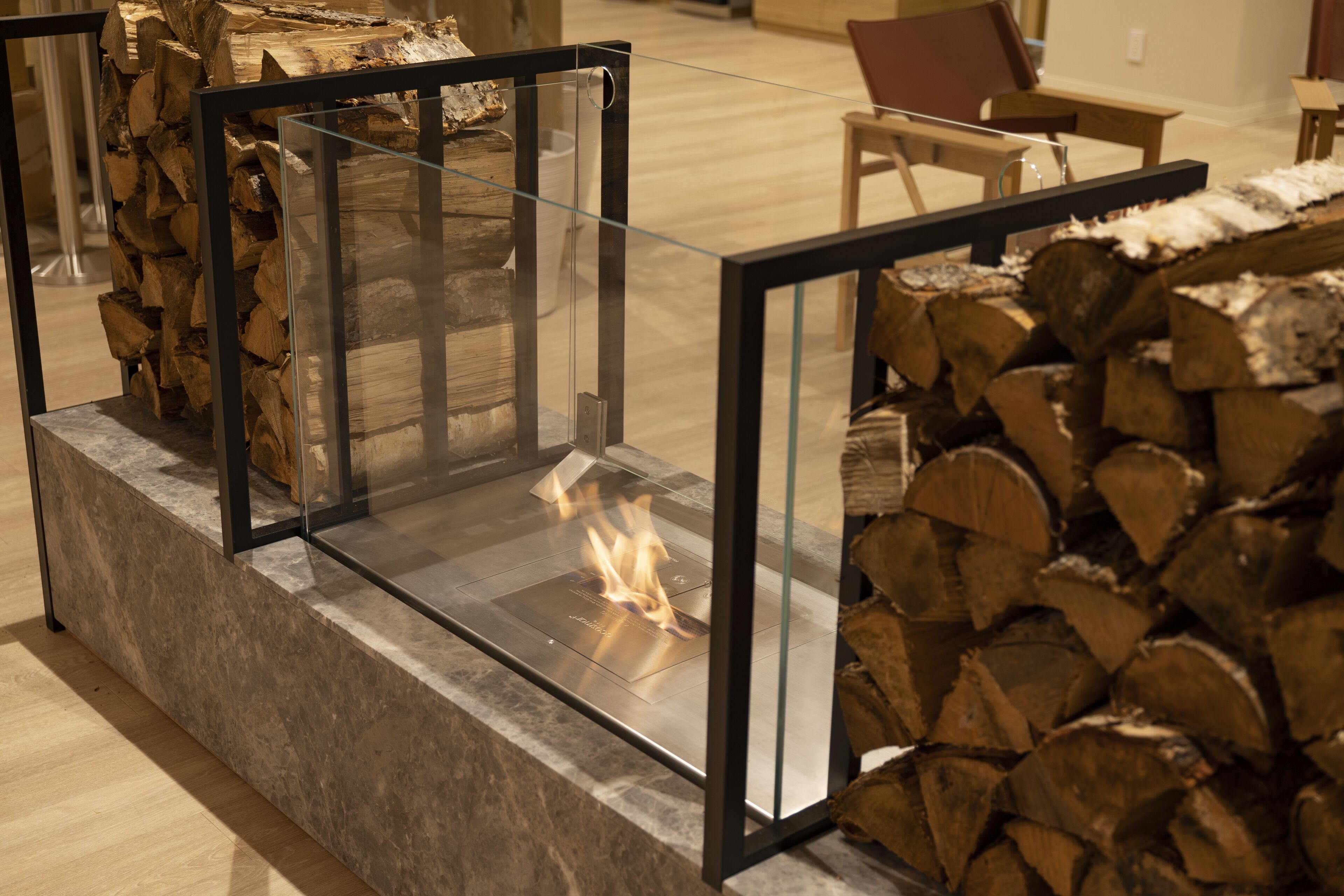 Ein von Glas umgebener Kamin mit sichtbarem Feuer, umgeben von gestapelten Holzscheiten.