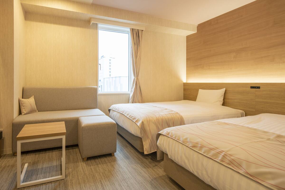 Une chambre d’hôtel avec un canapé, deux lits simples, une petite table et une fenêtre avec des rideaux.